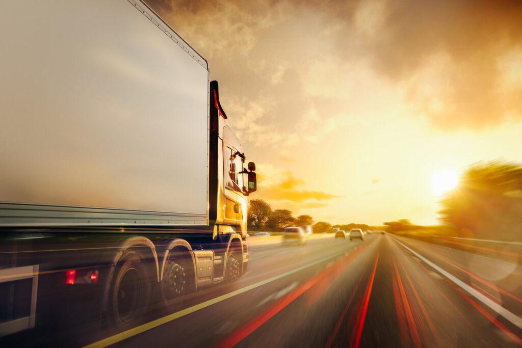 A side view of a moving semi-truck on a highway during sunset. The truck is in the right lane, and its motion is captured with a long exposure, creating light streaks on the road that convey a sense of speed. The sky is partly cloudy, and the warm hues of the setting sun fill the background, adding to the dynamic feel of the image