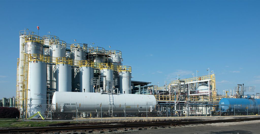 An industrial facility with multiple large storage tanks and complex piping. The tanks are cylindrical, with some standing vertically and others horizontally. The structure includes staircases, railings, and platforms for access. In the foreground, there are railway tracks suggesting transportation connectivity. The sky is clear blue, indicating good weather conditions