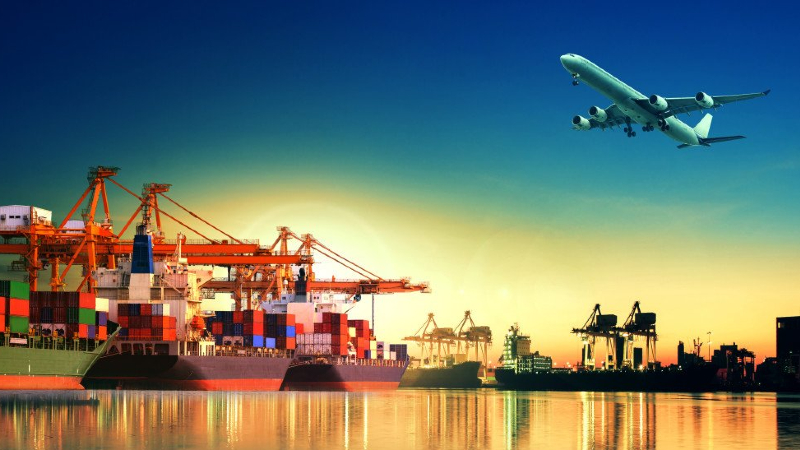 A commercial airplane taking off in a clear sky above a busy cargo port during sunset. The port is filled with colorful shipping containers stacked on large freight ships, and cranes are visible against the vibrant hues of the setting sun reflecting on the wate