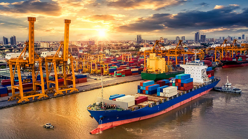 Aerial view of a busy shipping port at sunset. Multiple cranes tower over stacks of colorful shipping containers. A large cargo ship is docked, being loaded or unloaded, with a tugboat nearby on the water. The sky is painted with warm hues of orange and yellow from the setting sun, reflecting off the clouds and water, highlighting the industrial activity at the port