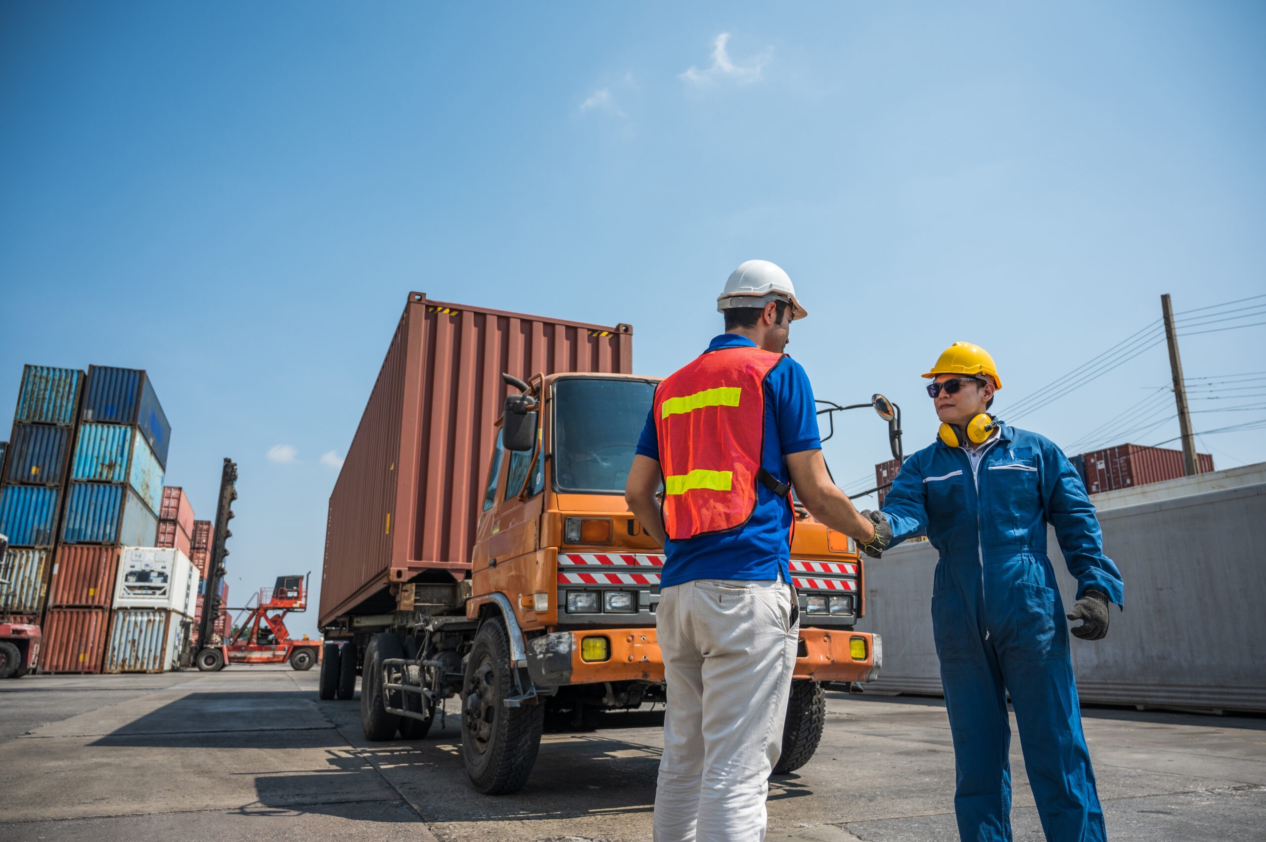 wo individuals in a shipping container yard during daytime. One person wears a white hard hat and a high-visibility vest, while the other operates a yellow device. Stacked shipping containers are visible in the background.